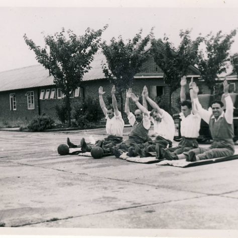 Stretching exercises outside the National Spinal Injuries Centre Black and white photo of five men sat in a row in on mats front of wheelchairs with arms stretched above their heads | Image courtesy of the National Paralympic Heritage Trust