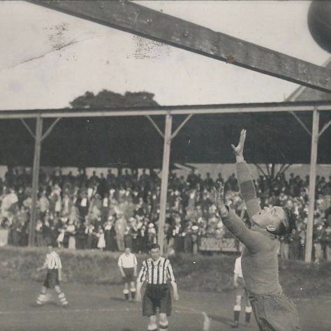HLF-Invite Black and white photo of a football goalkeeper diving for a ball which has travelled past the goalpost, as spectators and players watch on | Image courtesy of the Grecian Archive