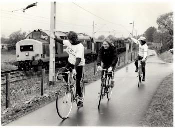 Image of three cyclists cycling along the Dumbarton Bike Path. They are waving at a train passing by. | West Dunbartonshire Council Archives