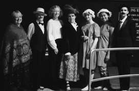 A black and white film still showing 7 members of the cast in Victorian style costumes
