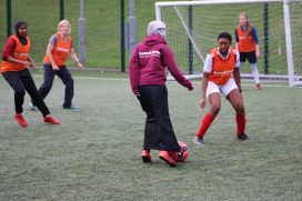 Woman wearing a FURD hoody and skirt running with ball towards defenders in front of goal.