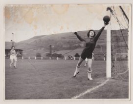 Female goalkeeper pushes ball away from the net, pennines in background to pitch