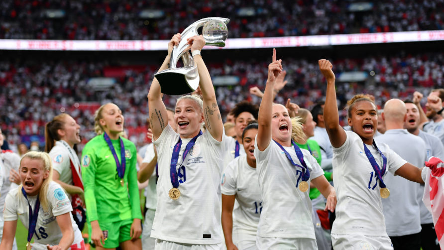 Women in white England kits raise trophy, dancing and cheering. Wembley crowd in background. | Colorsport / Ashley Western