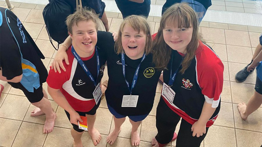 Three members of Down Syndrome Team in GB tshirts. Smiling at camera, arms around each others shoulders at poolside. | Down Syndrome Swimming Great Britain