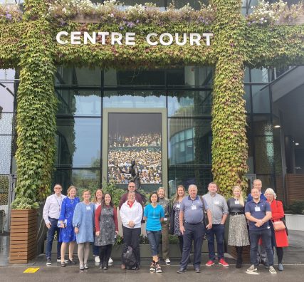 Group shot of attendees at the Sharing Stories event at Wimbledon standing in front of the Centre Court sign