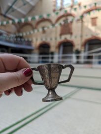 Hand holding small silver trophy, empty swimming baths behind.
