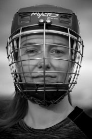 Close up of woman's face, wearing Shinty helmet with grilled visor down. In Black and white.