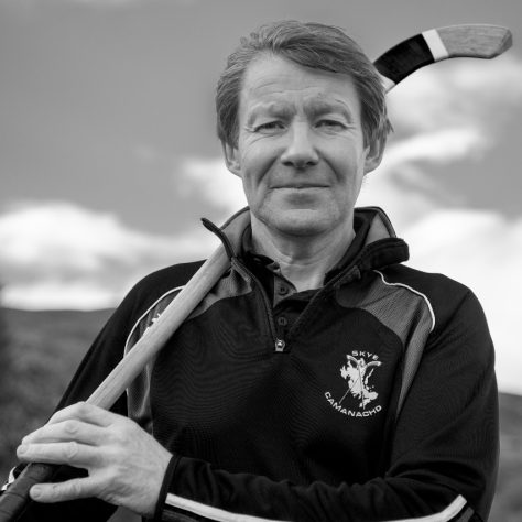Man wearing Skye Camanachd training top, holding a Shinty club over shoulder. In Black and White. | Isabelle Law Photography