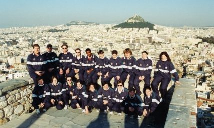 Cymru women wearing black tracksuits pose as team. Athens skyline and Mt Lycabettus in background