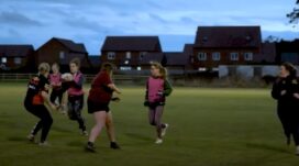 Six women play Rugby in coloured vests at night