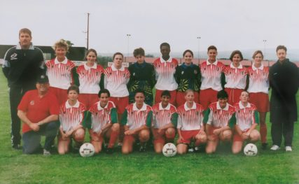 Women's team wearing red and white Wales kits pose together on pitch, alongside coaching staff.