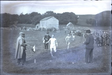 A black and white imageshowing a manabout to cross the finish line. He is in an all-whiteoutfit. There are other runners behind himand crowds on the right-handside. There are two men holding up the finish line. One is in a bowler hat and smart jacket and trousers. The other man is in a flat cap and long overcoat