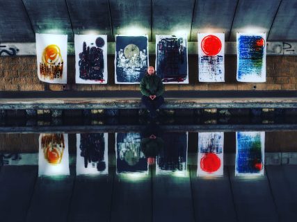 Man sits on canal side, six abstract art prints hang behind, reflecting in the water.