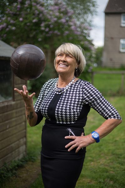 Rose Reilly balances a football in right hand, smiles with left hand on her hip. Wearing black and white dress, with short hair. Garden and shed in background.
