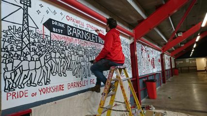 Ben sat on a ladder, paints Barnsley Mural wall panel in stadium corridor