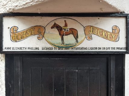 White Pub sign above black door showing illustrated horse and jockey, with scrolled title