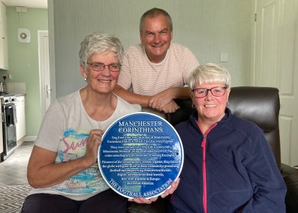 Two elderly women holding a blue plaque dedicated to a women’s football team with a man stood behind them