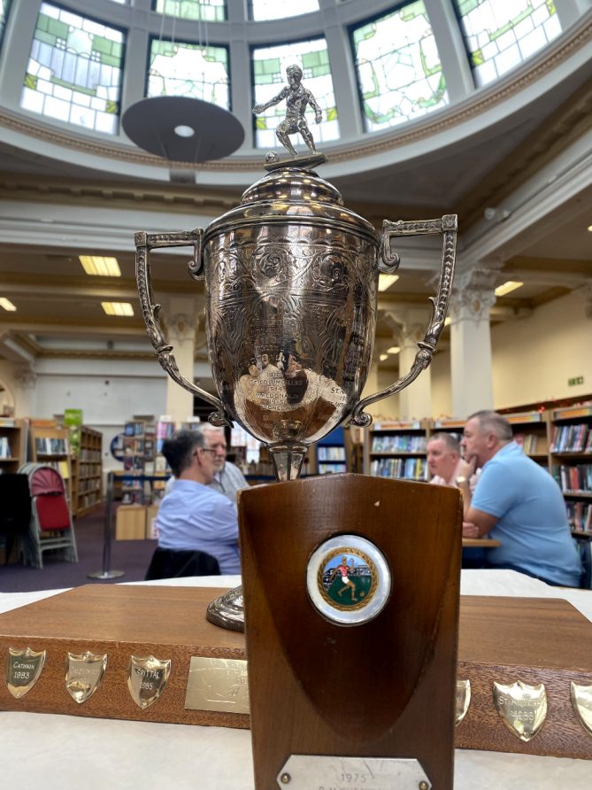 A silver sporting trophy with small wooden trophy in front of four seated men