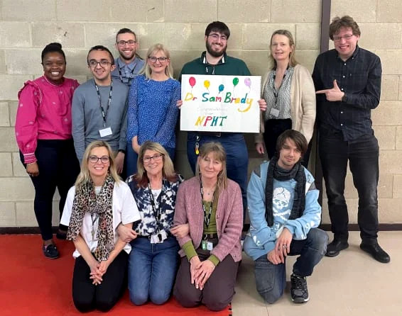 Team photo with Dr Sam Brady stood on back row holding colourful card with his name on.