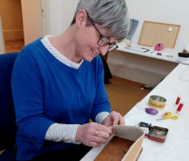 Colour photograph of woman wearing glasses with grey cropped hair and wearing a blue jumper sat at a desk using small hand tools to wotk on a museum object.