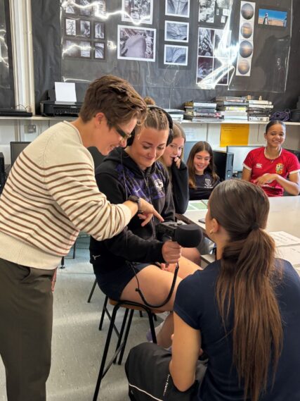 5 A group of students sit around a classroom table. One student wears headphones and holds a microphone while the workshop leader leans in to guide them. Others watch and smile. | © Sideline Assembly