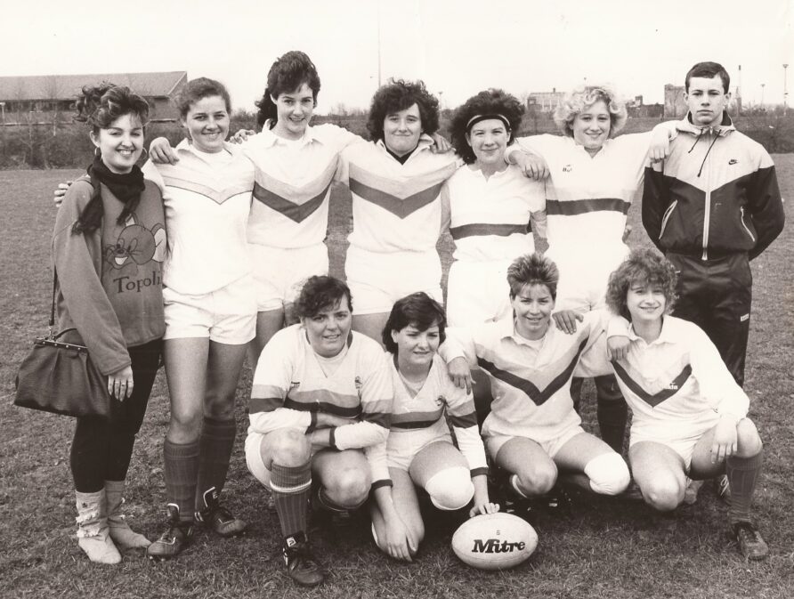Women in rugby strips pose for team photo, pitch in background